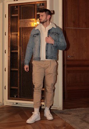 Chaqueta de mezclilla con cuello de borreguito, suéter blanco con cremallera, pantalones cargo beige y zapatillas blancas. Fondo de madera en tonos cálidos y ascensor vintage.