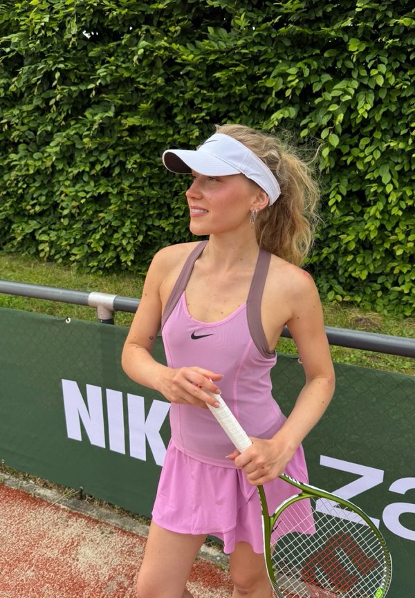 Jeune femme en tenue de tennis rose et visière blanche tenant une raquette de tennis, debout près d’un grillage avec de la verdure en arrière-plan.