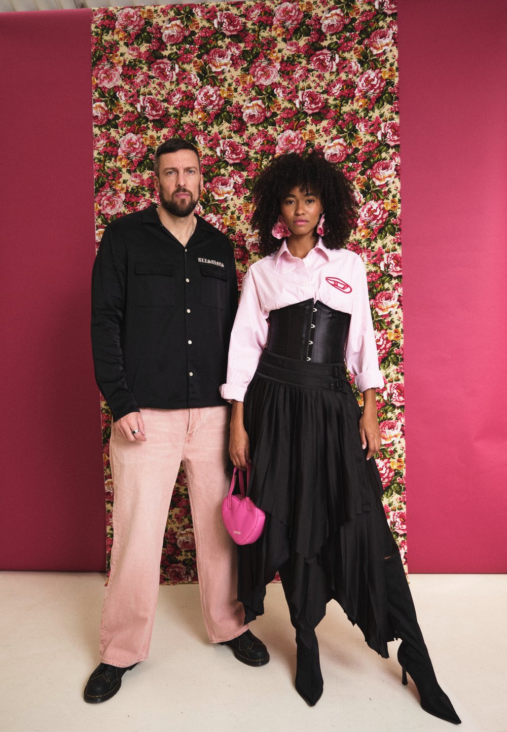 Camisa preta de botões, calças de veludo cotelê cor-de-rosa, camisa branca com riscas, corpete preto, saia preta em camadas, carteira cor-de-rosa, fundo floral.