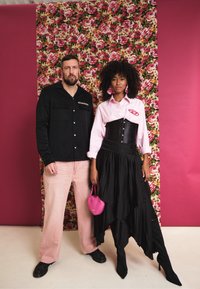 Camisa preta de botões, calças de veludo cotelê cor-de-rosa, camisa branca com riscas, corpete preto, saia preta em camadas, carteira cor-de-rosa, fundo floral.