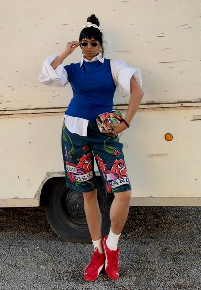 Mujer con gafas de sol, camisa blanca, top azul sin mangas, pantalones cortos estampados con flores, zapatillas deportivas rojas, sosteniendo un bolso de mano colorido, de pie junto a un vehículo blanco.