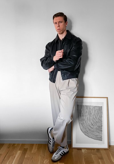 Joven con chaqueta de cuero negra, pantalones beige y zapatillas, apoyado contra una pared blanca junto a una obra de arte abstracta en blanco y negro enmarcada.