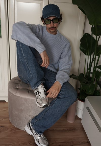Suéter acanalado de color azul claro, jeans de mezclilla azules, zapatillas grises y gafas de sol con tintado verde. Sentado en un ottoman redondo y suave de color gris claro.