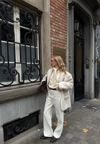 Blazer blanc texturé sur un pull crème et un pantalon large blanc, associé à des mocassins marron et un sac marron, devant un mur en briques.