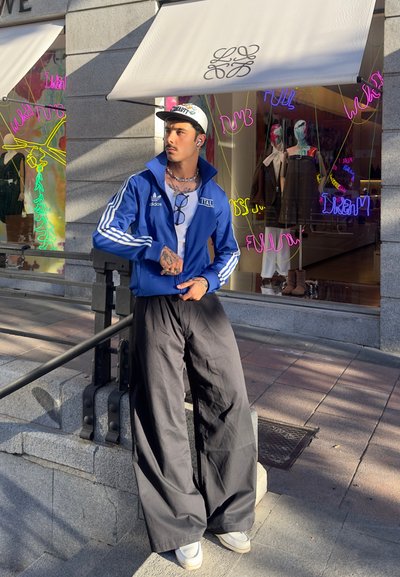 Chaqueta de chándal azul de Adidas con rayas blancas, pantalones anchos negros y zapatillas blancas, complementada con gafas y una gorra, de pie frente a una tienda.