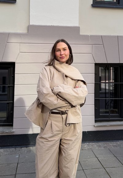 Mujer con chaqueta y pantalones beige de gran tamaño, con los brazos cruzados, apoyada contra una pared de piedra clara con ventanas con marcos negros al aire libre.