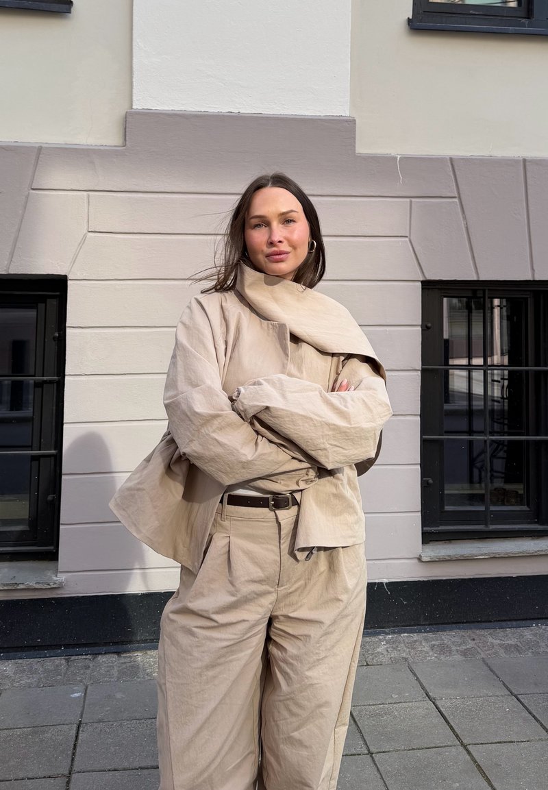 Woman in beige oversized jacket and pants stands with arms crossed against a light stone wall with black-framed windows outdoors.