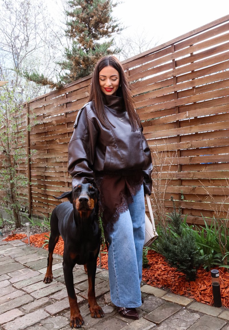 Frau mit langen dunklen Haaren, die eine braune Lederjacke und blaue Jeans trägt, steht neben einem schwarzen Dobermann auf einem Steinweg neben einem Holzzaun.