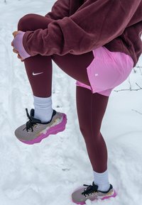 Personne en leggings bordeaux, shorts roses et veste violette s'étirant une jambe tout en se tenant debout sur un sol enneigé, portant des chaussures de course grises et roses.