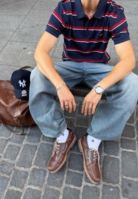 Kék és piros csíkos pólót viselő személy, világoskék farmerben, barna bőrcipőben, ezüst órával és gyűrűkkel ül egy térköves burkolatú kőn, közel egy táskához és sötétkék sapkához.