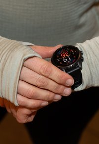 Rankos reguliuoja juodą išmanųjį laikrodį, rodomą laiką, datą ir fizinius rodiklius, užsiėmus šviesios spalvos ilgomis rankovėmis.