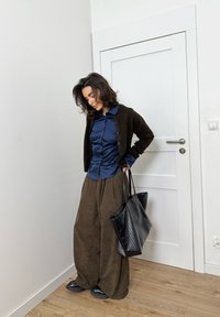 Barna bőszárú nadrág, tengerészkék szatén gombos ing, fekete kötött kardigán és texturált fekete válltáska. Semleges háttér és fa padló.