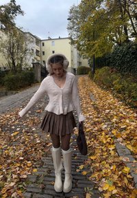 Creme strikket cardigan, brun plisseret nederdel, creme knæhøje støvler og beige ørevarmere. Efterårsblade på en brostensbelagt sti i baggrunden.