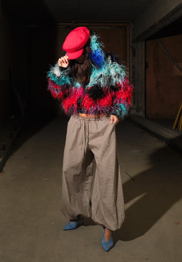 Person wearing a colorful furry jacket, red cap, checkered wide pants, and blue pointed heels standing against a dark background.