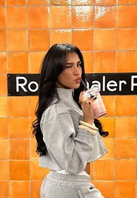 Šedá zkrácená mikina a odpovídající kalhoty. Dlouhé tmavé vlasy, držící růžový smoothie v průhledném hrnku s černým brčkem. Oranžová dlaždicová pozad�í.