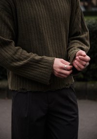 Pull en tricot côtelé vert olive avec un ourlet brut et des manches larges, associé à un pantalon sombre sur mesure. Anneaux visibles aux mains.