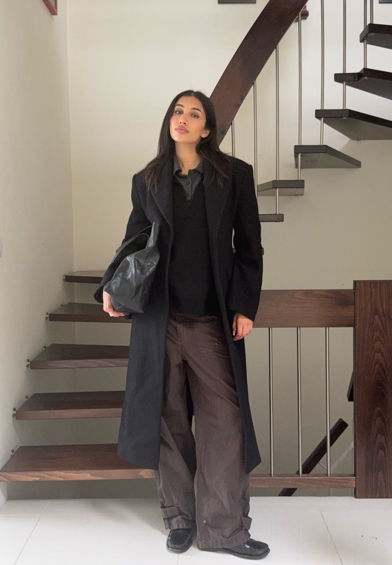 Cappotto lungo nero sopra una maglia scura, abbinato a pantaloni marroni a gamba larga. Tiene in mano una borsa nera texturizzata, con una scala di legno sullo sfondo.