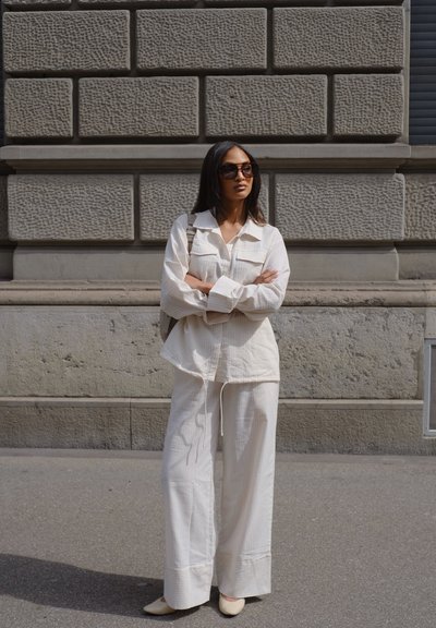 Camisa de gran tamaño en color crema y texturizada, y pantalones de pierna ancha con sutiles rayas. Complementados con gafas de sol oscuras y zapatos beige.