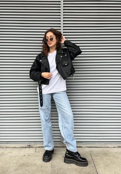 Chaqueta de cuero negra corta con cremalleras, camiseta blanca, pantalones vaqueros de pierna ancha azul claro y botas de plataforma negras gruesas. Gafas de sol circulares.