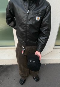 Zwarte leren jas met een kraag, ritssluiting en embleem. Bruine gestreepte broek, glanzende zwarte schoenen en een zwarte pet in de hand, met tekst.