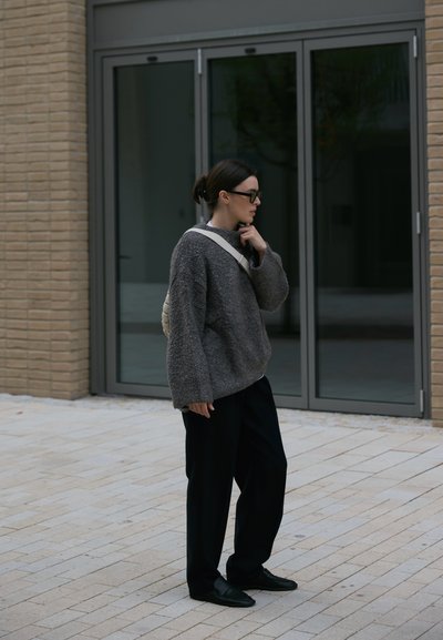 Suéter gris de gran tamaño y textura, combinado con pantalones de pierna ancha oscuros y zapatos negros. Una mochila blanca y unas gafas completan el conjunto.