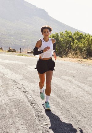 Corridore atletico su una strada, indossa una maglietta bianca senza maniche, pantaloni neri, maniche per le braccia e scarpe da corsa turchesi. Sfondo montano.