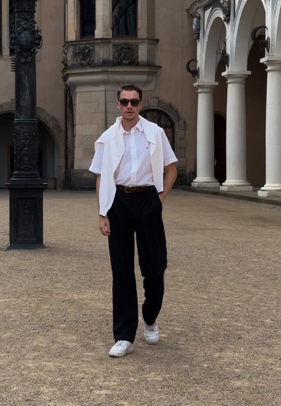 Camisa blanca, pantalones de rayas negras, zapatillas blancas, gafas de sol y un suéter blanco colgado sobre los hombros, de pie en un patio.