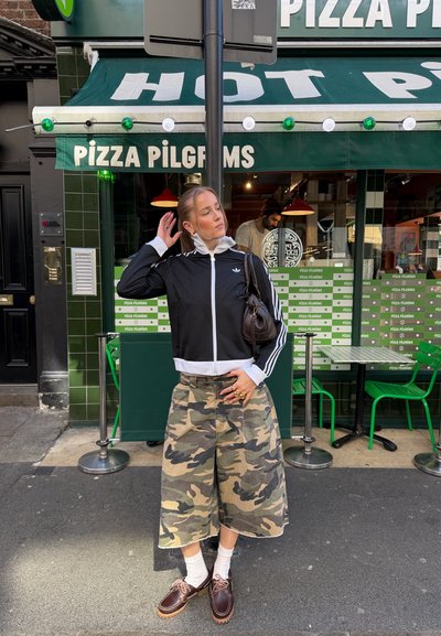 Mujer con falda de camuflaje y chaqueta negra está delante de Pizza Pilgrims, con sillas y mesas verdes visibles en el interior.