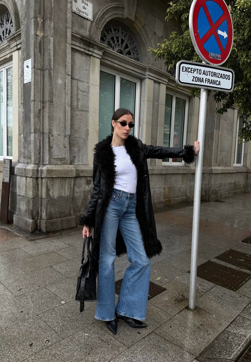 Donna con cappotto nero rifinito in pelliccia, top bianco, jeans a gamba larga e stivali neri, appoggiata a un palo metallico con un cartello di divieto di sosta.