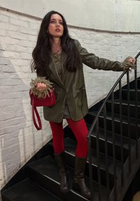 Blazer de cuero verde oliva sobre una blusa beige texturizada, combinado con medias rojas y botas marrones hasta la rodilla. Pequeño bolso rojo con correa para el hombro.