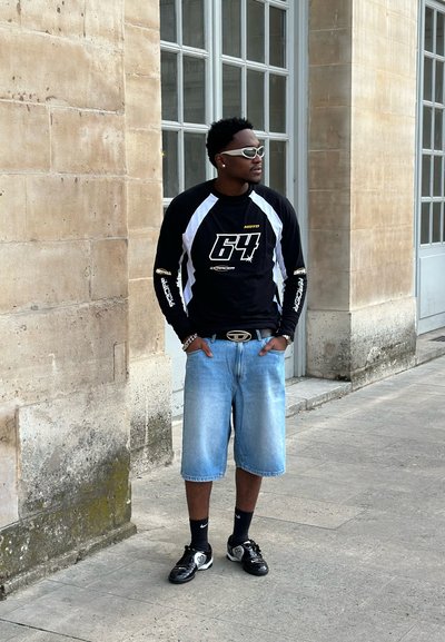 Camisa negra de manga larga con acentos blancos y un gran logo "64", combinada con pantalones cortos de mezclilla azul claro y zapatillas deportivas negras. Gafas de sol blancas.