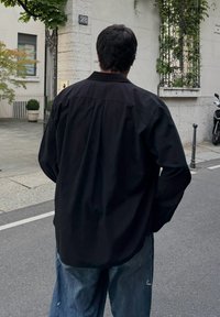 Černá košile na knoflíky s volným střihem a zadním dílem se stojkou. Nošena s světlými modrými roztrhanými volnými džíny na městské ulici.