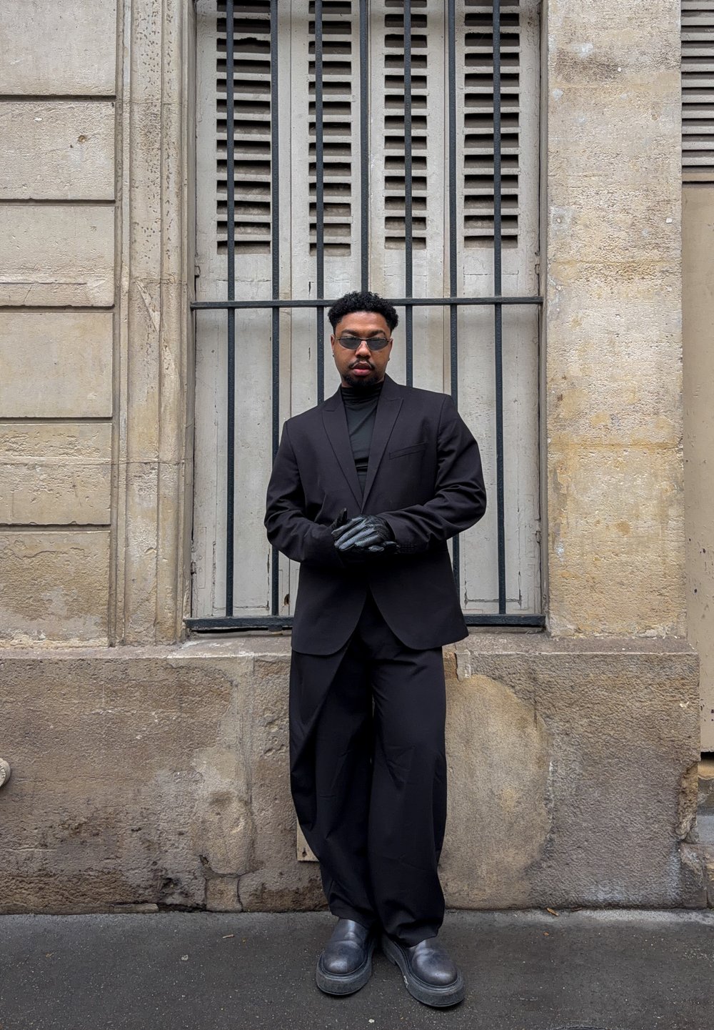 Veste noire cintrée portée sur un col roulé noir, associée à un pantalon large noir et des chaussures noires épaisses. Porte des lunettes de soleil et des gants noirs.