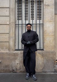 Veste noire cintrée portée sur un col roulé noir, associée à un pantalon large noir et des chaussures noires épaisses. Porte des lunettes de soleil et des gants noirs.