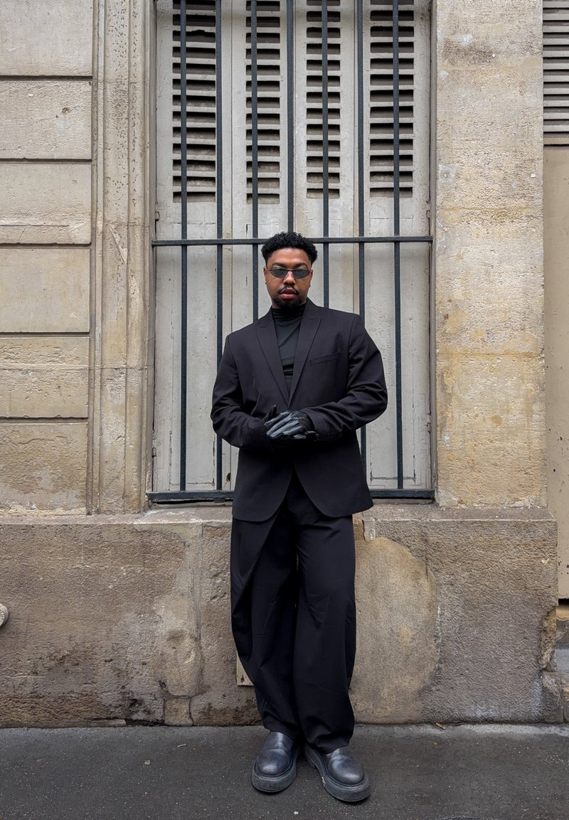 Schwarzer maßgeschneiderter Blazer über einem schwarzen Rollkragenpullover, kombiniert mit weiten schwarzen Hosen und wuchtigen schwarzen Schuhen. Trägt Sonnenbrille und schwarze Handschuhe.