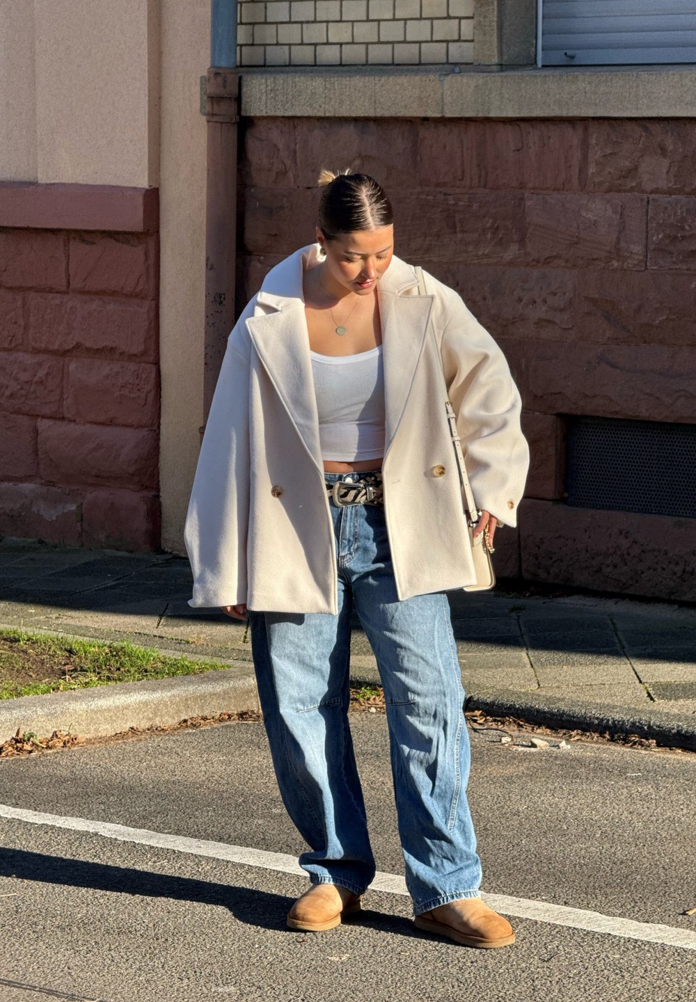 Casaco oversized creme, top cropped branco, jeans baggy azul claro e sapatos slip-on bege; detalhes notáveis incluem um cinto prateado e uma bolsa.