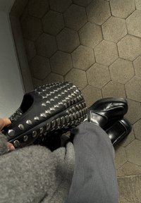 Mala preta com tachas metálicas, superfície texturizada, segurada numa mão. Sapatos de couro pretos com biqueiras arredondadas, calças cinzentas, chão com azulejos hexagonais.