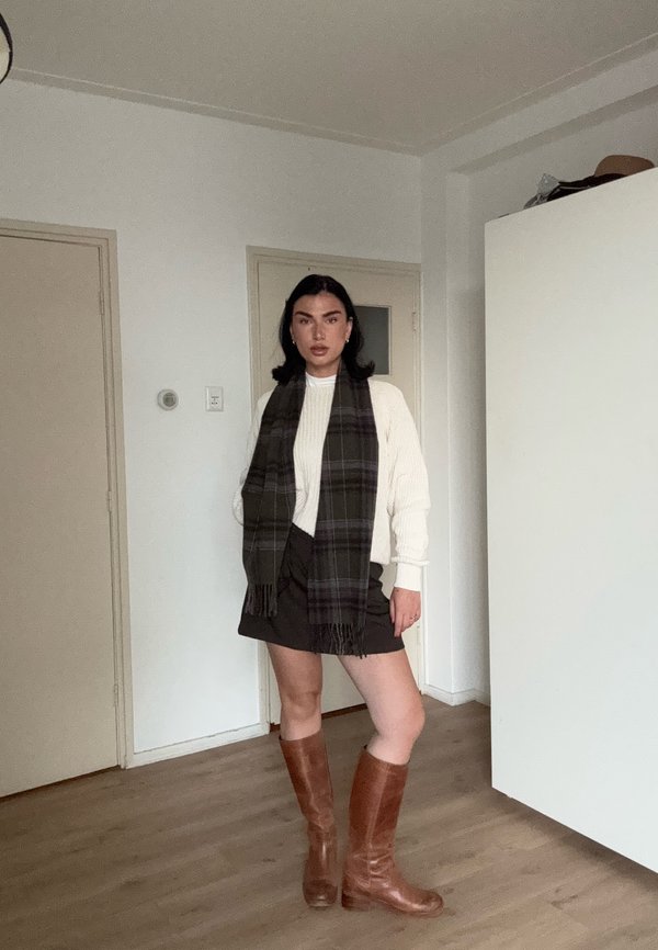Una mujer luce en una habitación un jersey blanco, una falda negra, una bufanda larga de cuadros oscuros y unas botas marrones hasta la rodilla.
