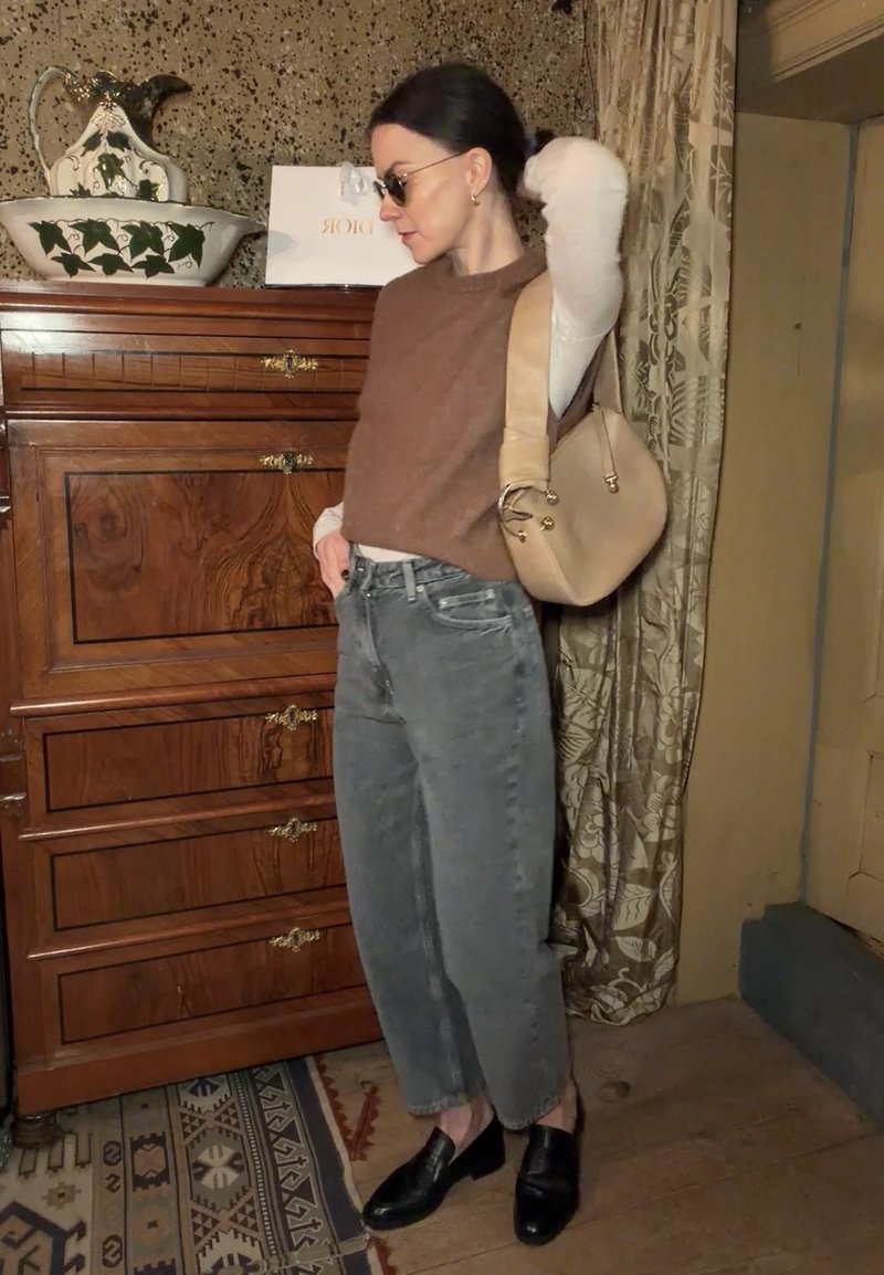 Kvinde med solbriller iført en brun ærmeløs sweater, hvid langærmet skjorte, grå jeans, sorte sko og holder en beige håndtaske indendørs.