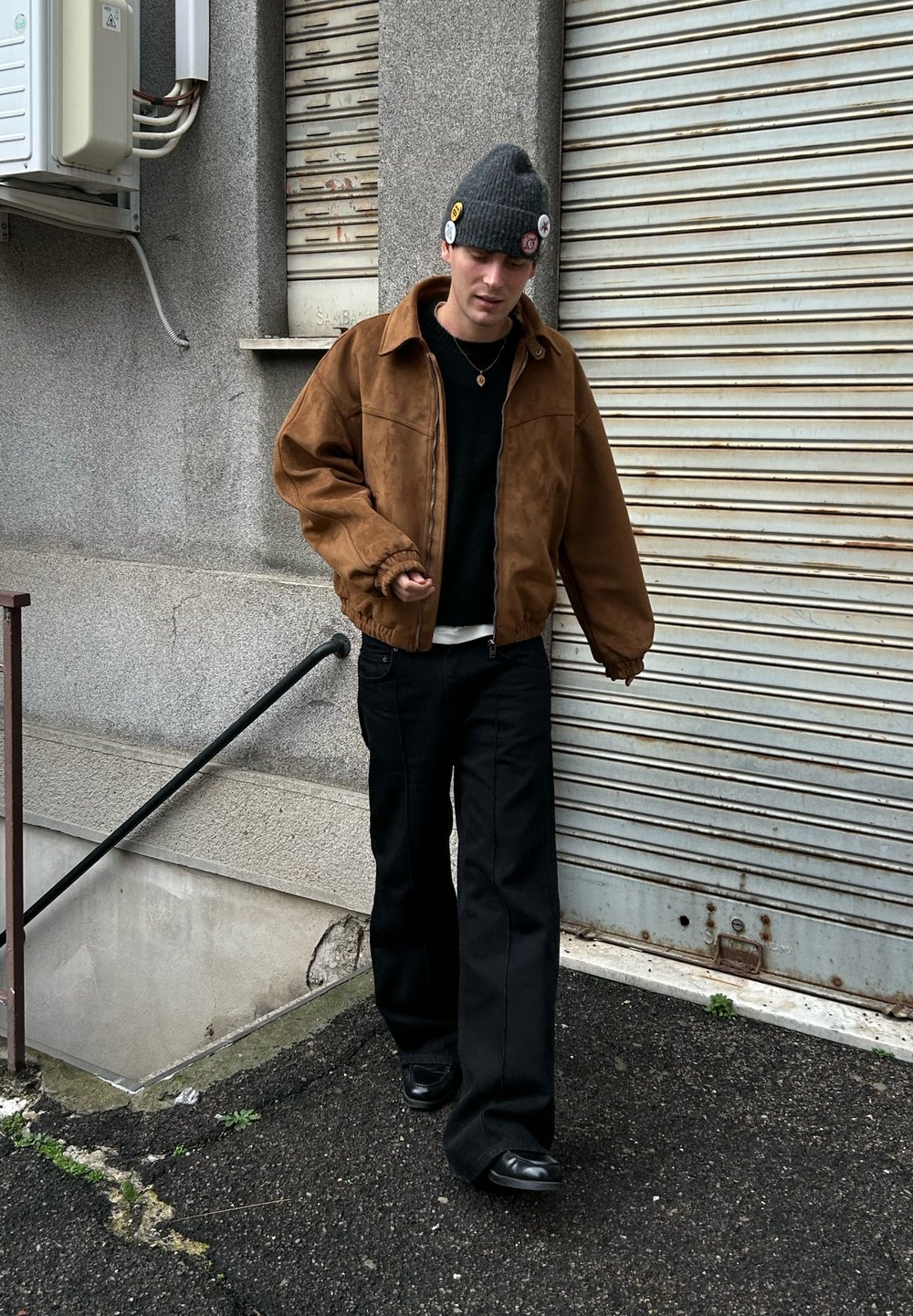 Blouson aviateur en daim marron, pull noir, pantalon large noir et chaussures noires. Bonnet en tricot gris avec des boutons. Cadre urbain.