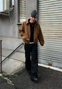 Blouson aviateur en daim marron, pull noir, pantalon large noir et chaussures noires. Bonnet en tricot gris avec des boutons. Cadre urbain.
