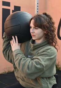 Schwarzer Gymnastikball, der von einer Person in einer grünen Fleece-Haube über der Schulter gehalten wird. Im Hintergrund ist eine strukturierte pinkfarbene Wand mit schwarzen Akzenten zu sehen.