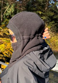 Mustast fliisist kapuuts, millel on nööriekra, kantud tumeda jope peal, tihedalt istuv ja kantud toonitud päikeseprillidega.