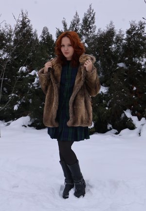 Mujer de cabello rojo con abrigo de piel, vestido de cuadros, medias negras y botas, de pie en la nieve con árboles perennes de fondo.