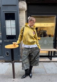 Hoodie jaune à fermeture éclair avec logo, chemise blanche à col, shorts cargo camouflage, et bottes noires pointues montant jusqu'aux genoux, tenant une boisson glacée sur une table en bois.