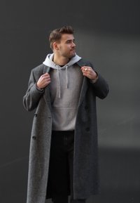 Szary wełniany płaszcz o teksturowanym wykończeniu, noszony na jasnoszarym hoodie. Posiada podwójnie zapinane guziki i luźny krój, w połączeniu z ciemnymi dżinsami.