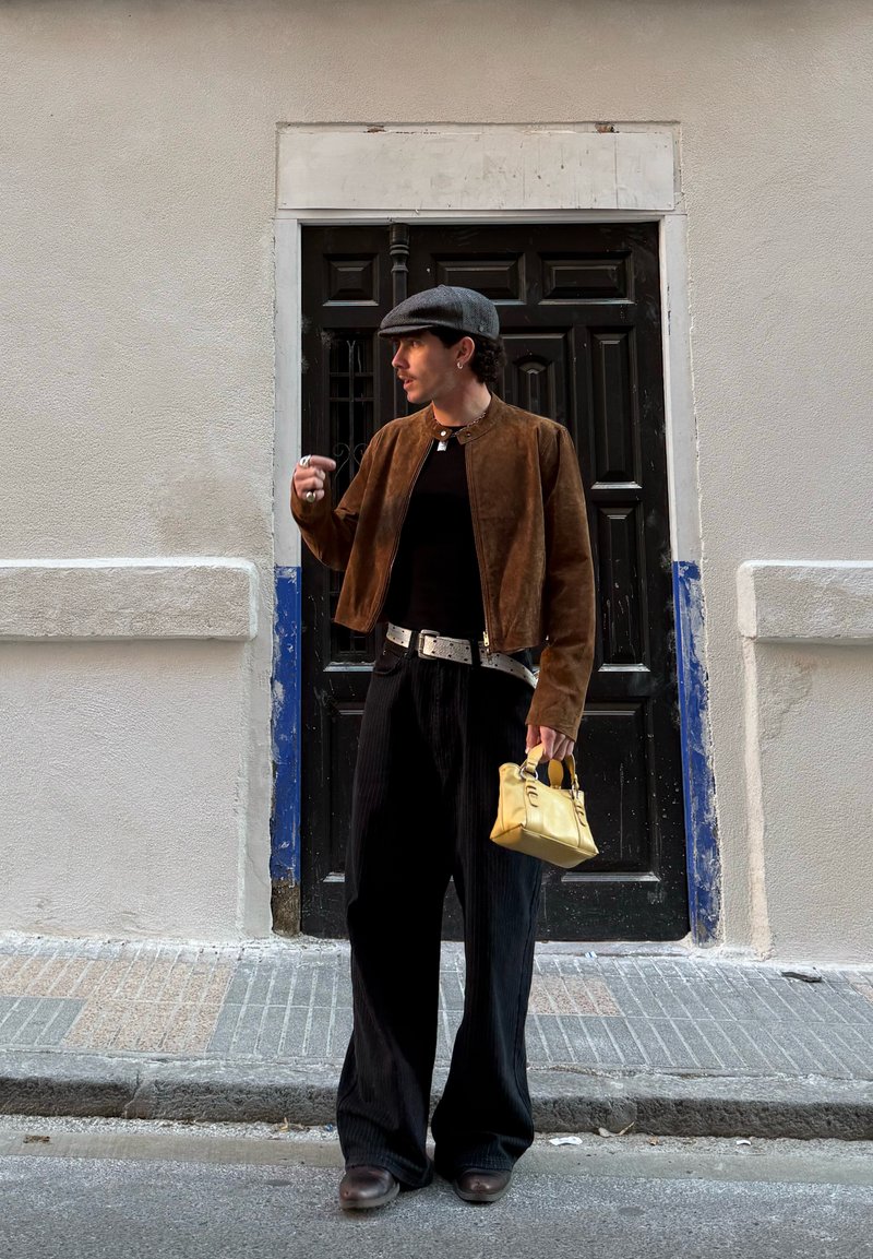 Personne portant une veste marron et une casquette plate se tient devant une porte noire, tenant un petit sac à main jaune et regardant sur le côté.