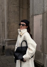 Valkoinen villakangastakki korkealla kauluksella ja mustilla napeilla, yhdistettynä mustaan käsilaukkuun. Lisäksi musta baretti ja aurinkolasit täydentävät asun.