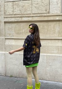 Schwarzes Trikot mit Diamantmuster und goldener Nummer 24, kombiniert mit einem leuchtend grünen Minirock und gelben knielangen Stiefeln. Gelbe Sonnenbrille.
