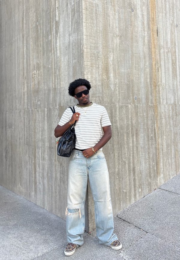 Hombre con gafas de sol, camiseta de rayas, vaqueros anchos rotos y zapatos con estampado, está apoyado contra una alta pared de esquina de hormigón sosteniendo una bolsa negra.